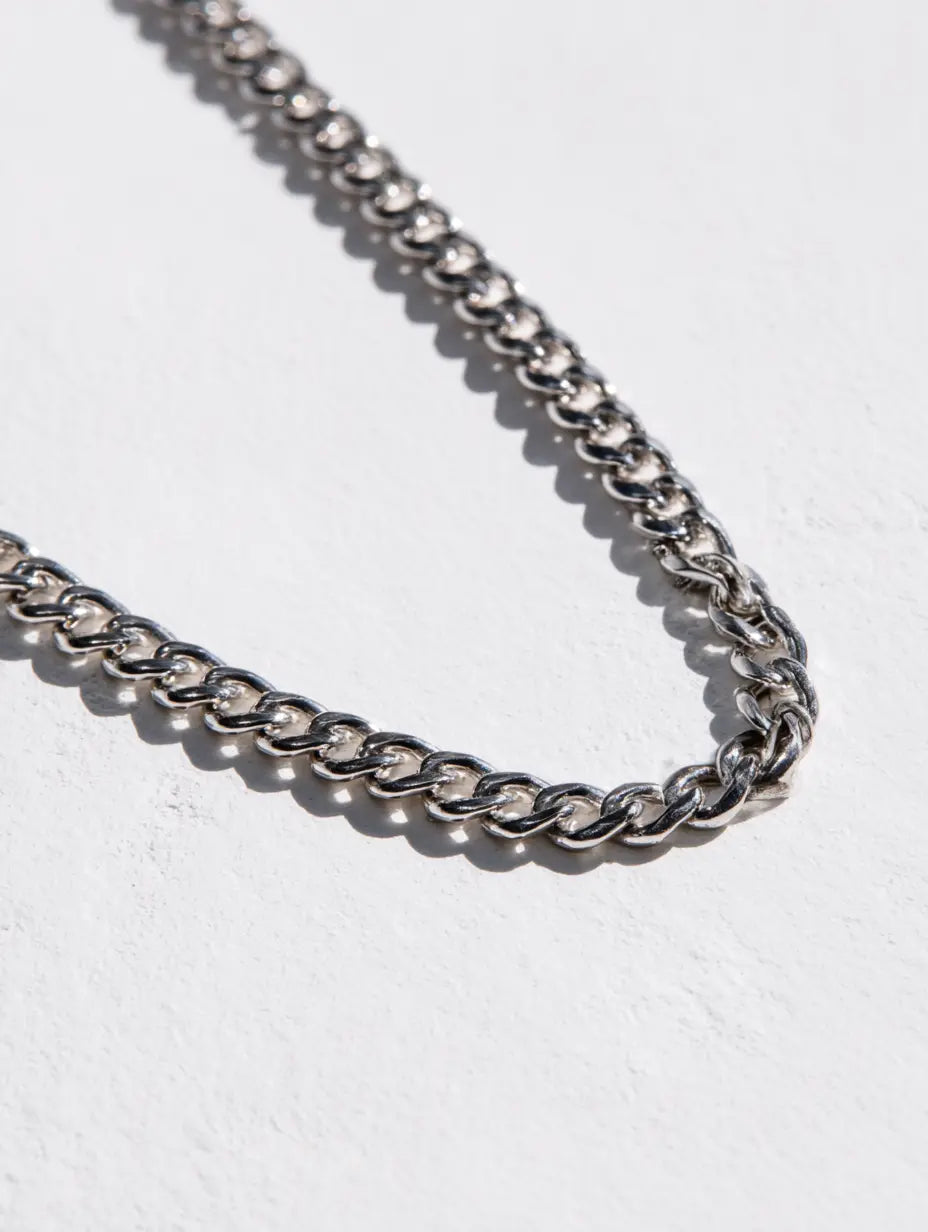A close-up of a silver-toned cuban chain necklace  by Ravion draped diagonally across a textured white surface.