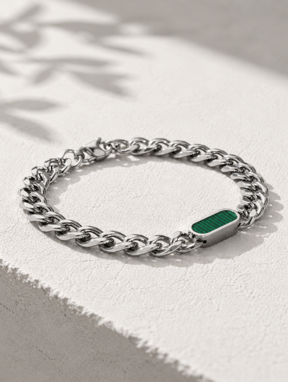 A silver-toned curb chain bracelet with a polished rectangular green inlay, displayed on a textured white surface with soft leaf shadows.
