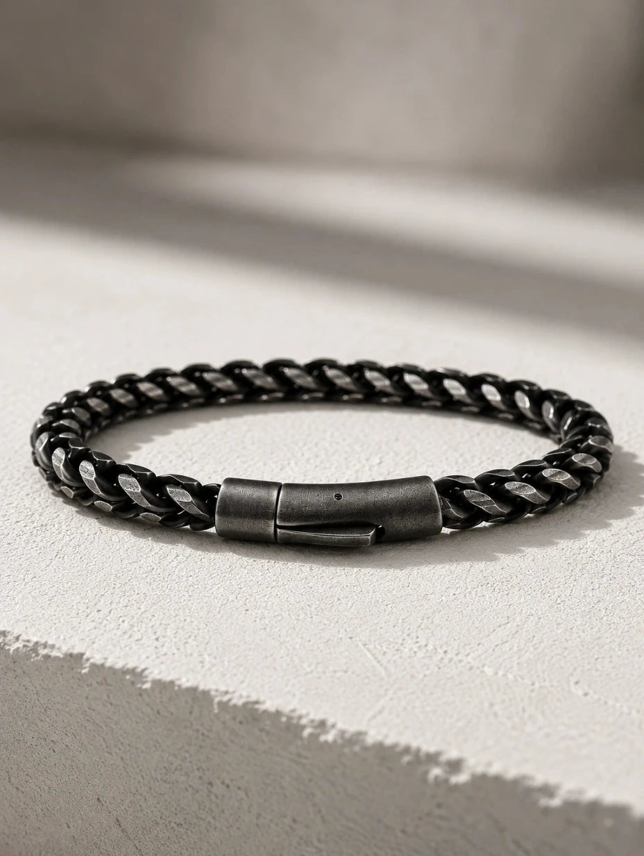 A black braided leather bracelet with a dark gunmetal metal clasp, resting on a textured light gray surface with soft shadows.