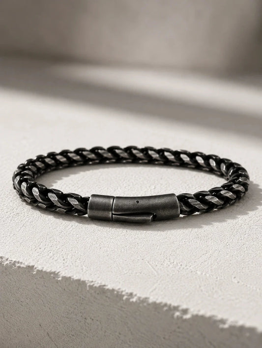 A black braided leather bracelet with a dark gunmetal metal clasp, resting on a textured light gray surface with soft shadows.