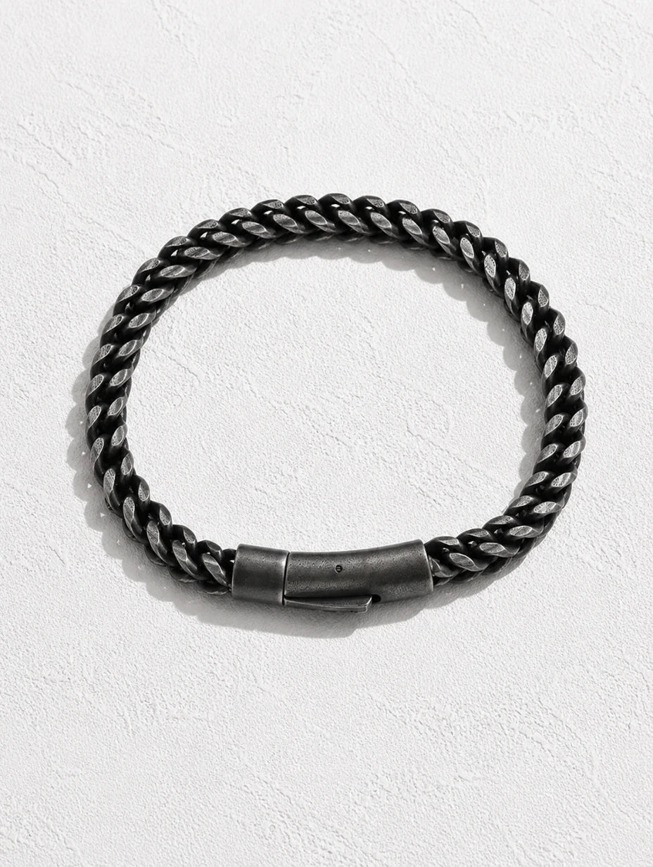 A thick braided rope-style bracelet in black and silver tones with a dark metal clasp, displayed on a textured white background.