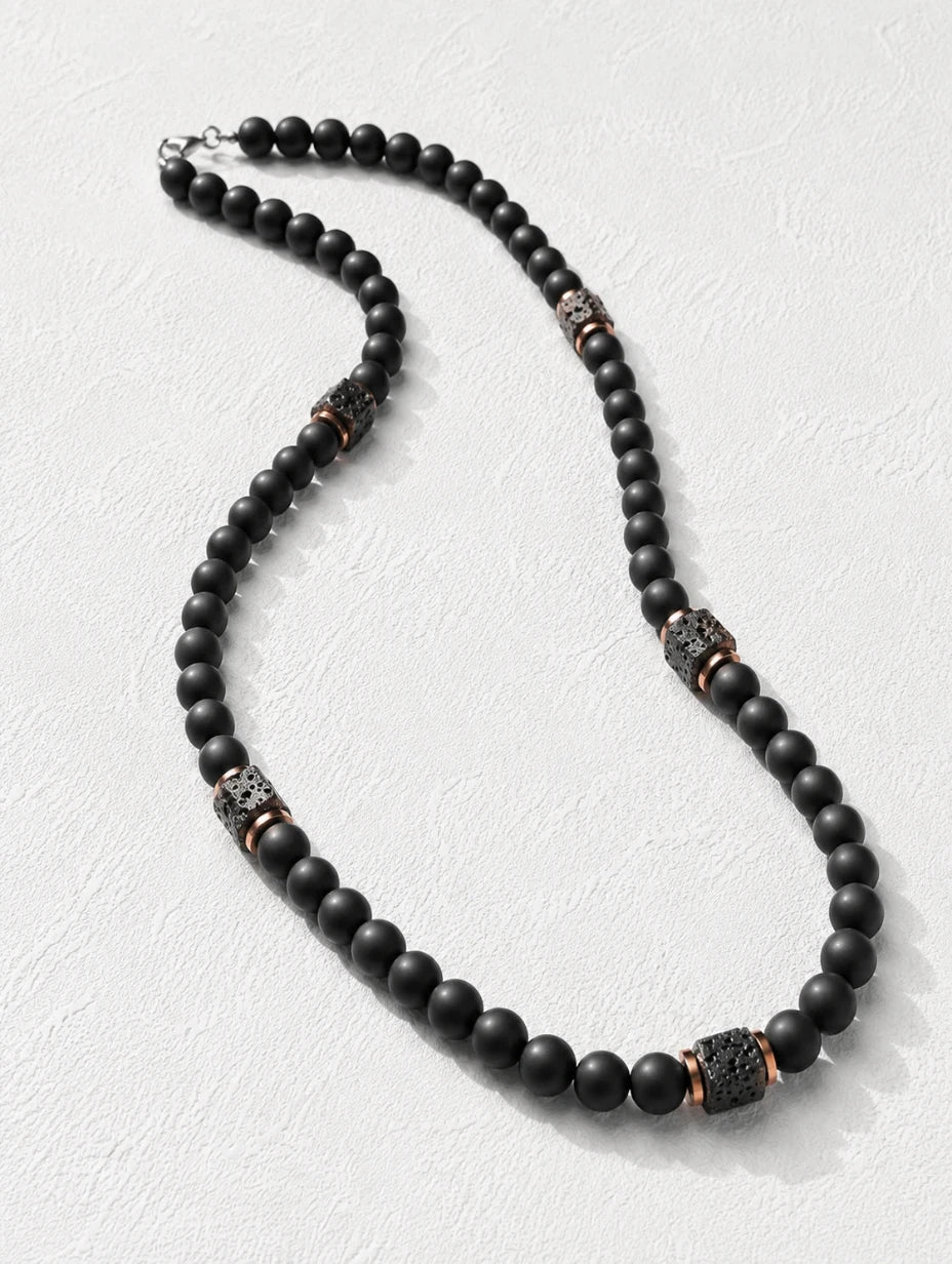 The Obsidian  beaded necklace featuring matte black round beads with textured metallic spacer beads in a bronze and silver finish, displayed on a light textured background.