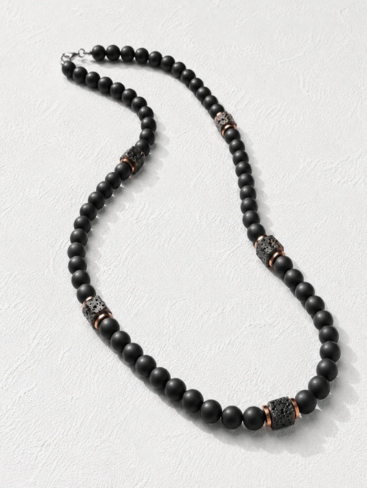 The Obsidian  beaded necklace featuring matte black round beads with textured metallic spacer beads in a bronze and silver finish, displayed on a light textured background.