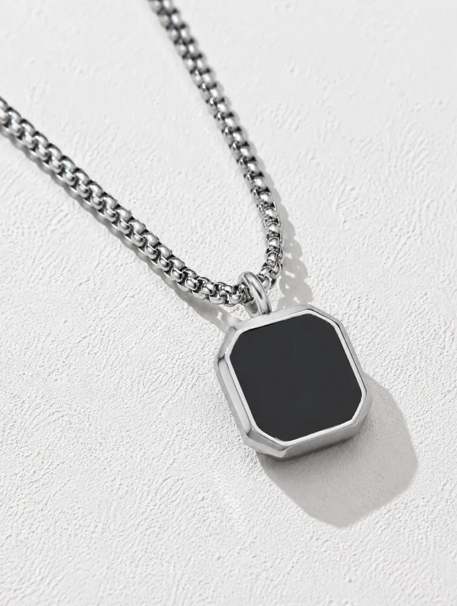 A silver-toned chain necklace with a faceted octagonal pendant featuring a matte black center, displayed on a textured light gray background.