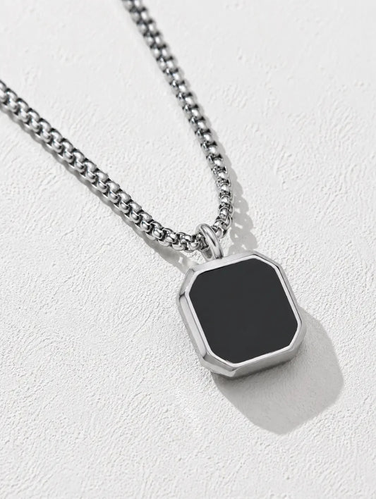 A silver-toned chain necklace with a faceted octagonal pendant featuring a matte black center, displayed on a textured light gray background.