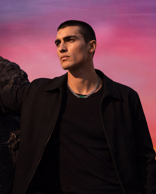 A man wearing a black jacket and black t-shirt, showcasing a silver chain necklace with a Ravion meridian pendant necklace posed against a vibrant pink and purple sky backdrop.